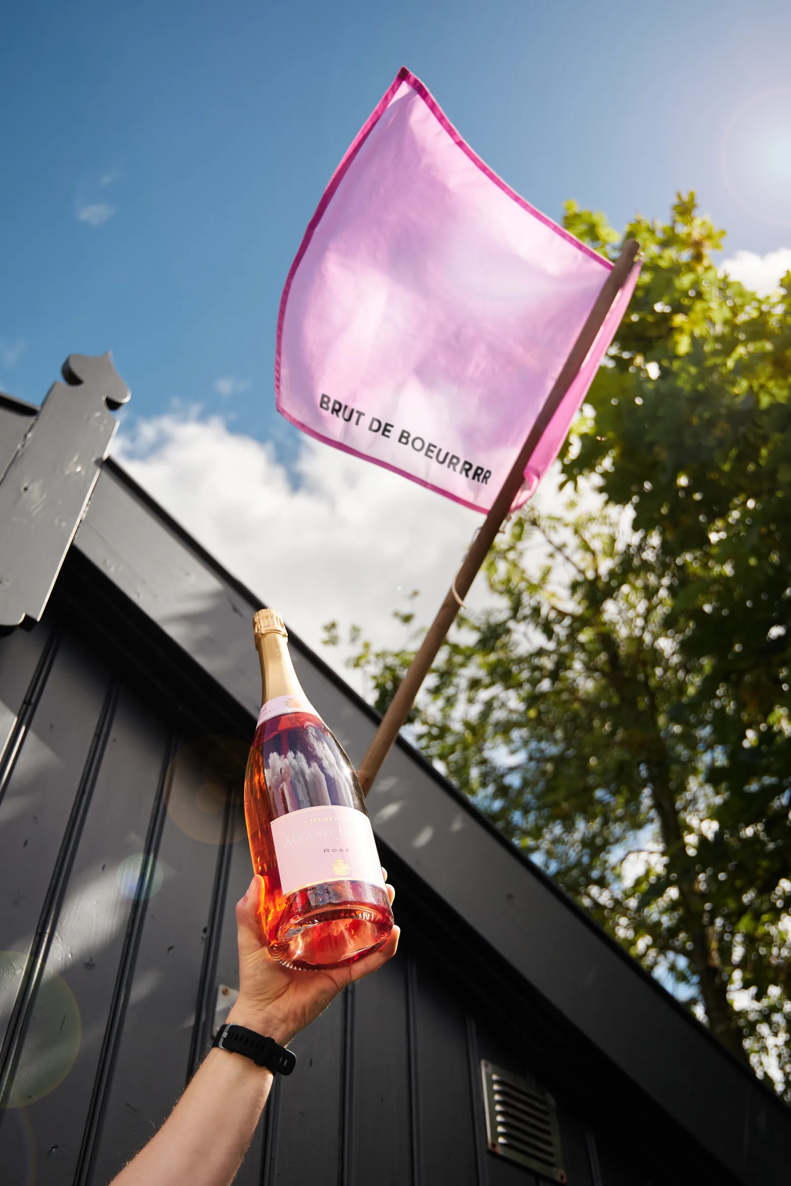 Hand met een fles rosé champagne en roze Brut de Boeurrrr-vlag tegen een blauwe zomerlucht